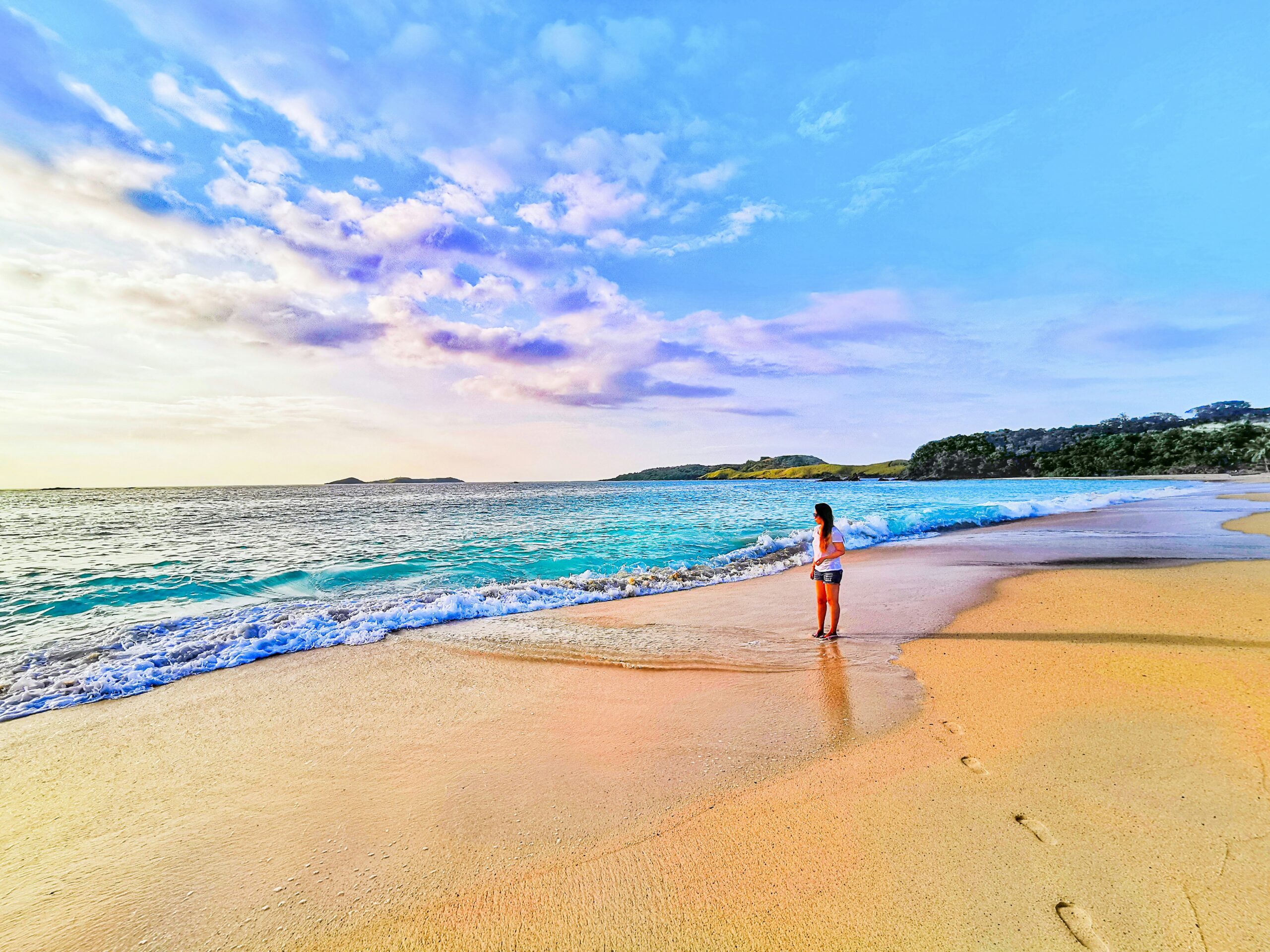 A woman stands on the sandy shore of Calaguas Island in Camarines Norte, Philippines, gazing at the vibrant blue ocean under a sky painted with soft clouds at sunset.