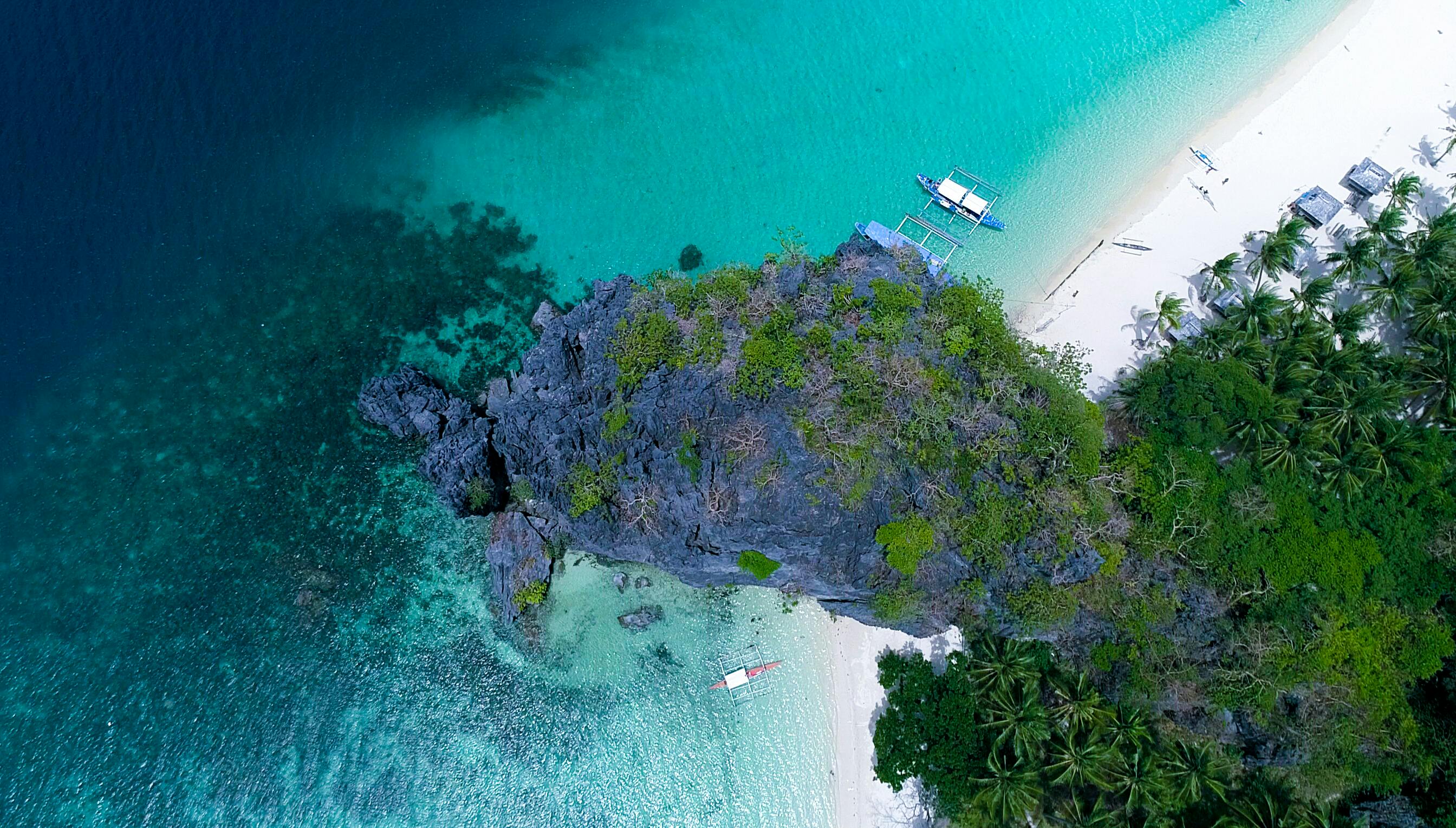 An aerial view of a pristine beach in El Nido, featuring a dramatic rocky outcrop covered in greenery, surrounded by turquoise waters and coral reefs, with traditional Filipino outrigger boats (bancas) anchored near the white sandy shore and lush palm trees lining the beach.