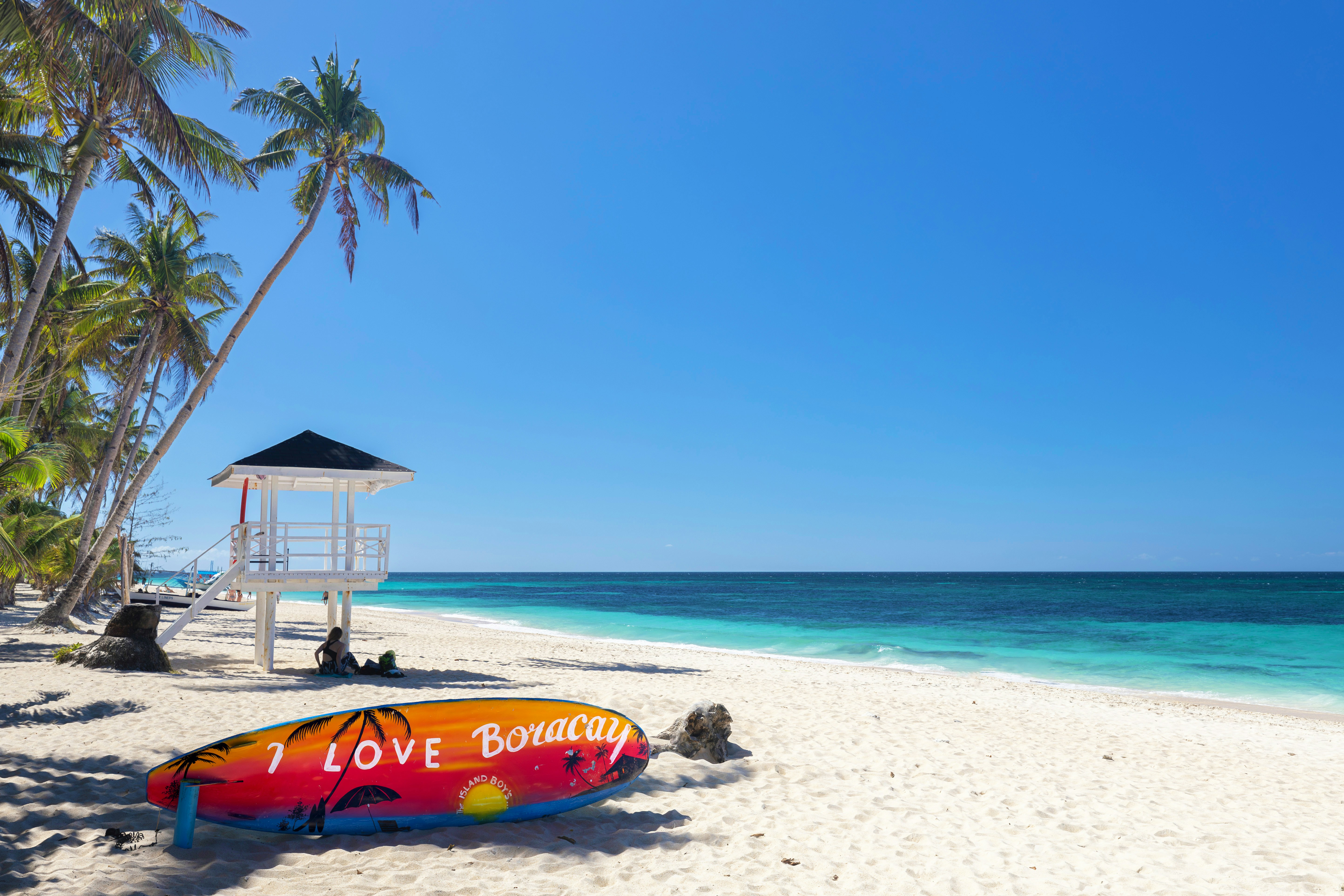 A tranquil beach scene in Boracay with clear blue skies, turquoise waters, a lifeguard tower, and a vibrant surfboard painted with "I Love Boracay."