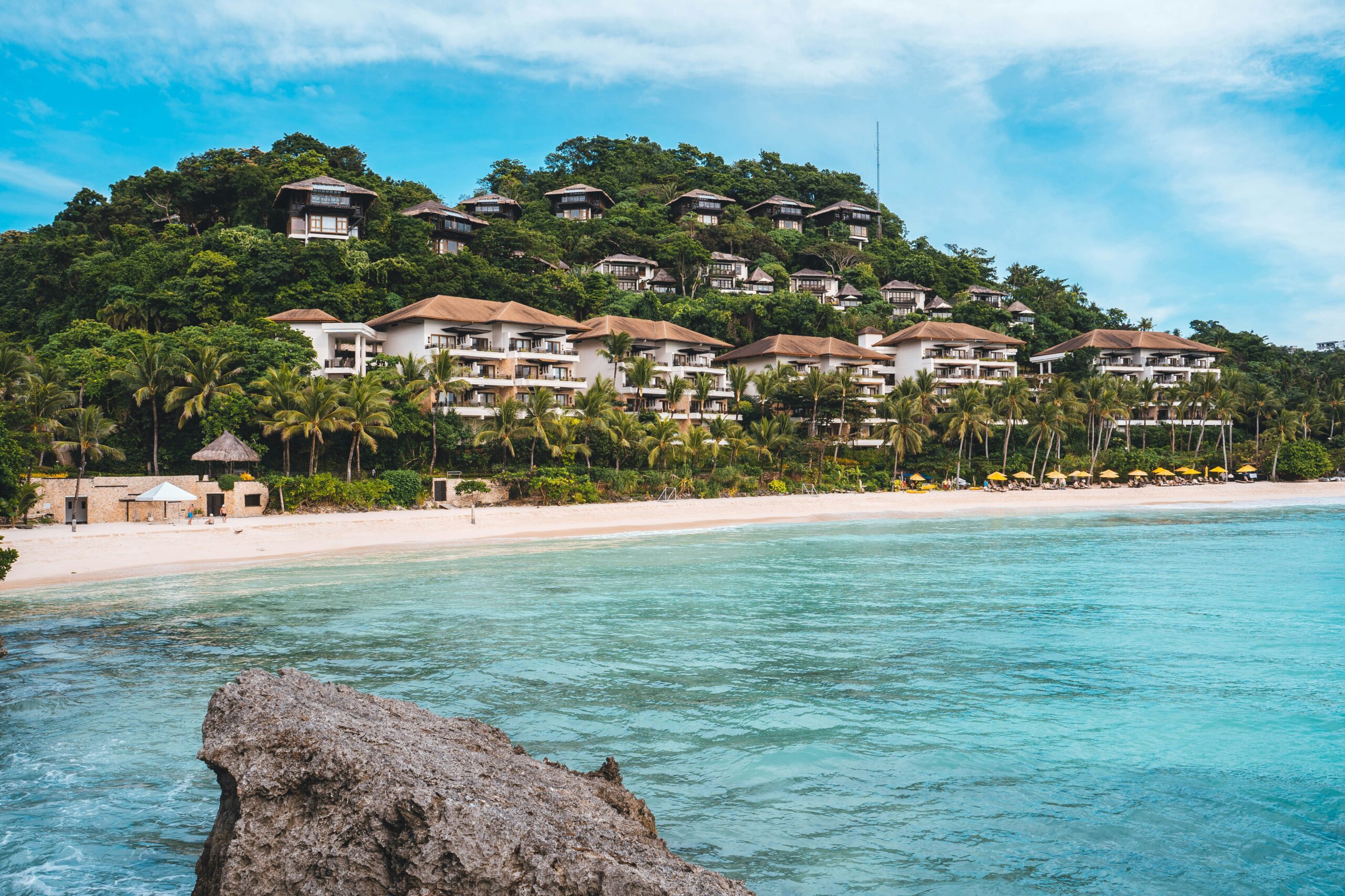 A tropical beach resort with modern and traditional architecture sits nestled in lush green hills, overlooking a pristine white sand beach and turquoise waters, lined with palm trees and beach umbrellas.