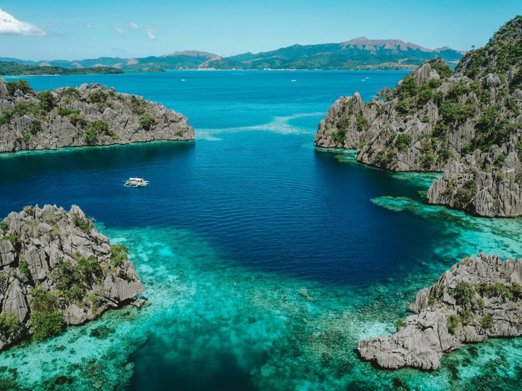 Aerial view of Coron, Palawan, featuring clear turquoise waters, dramatic limestone cliffs, and a traditional Filipino boat in the center.