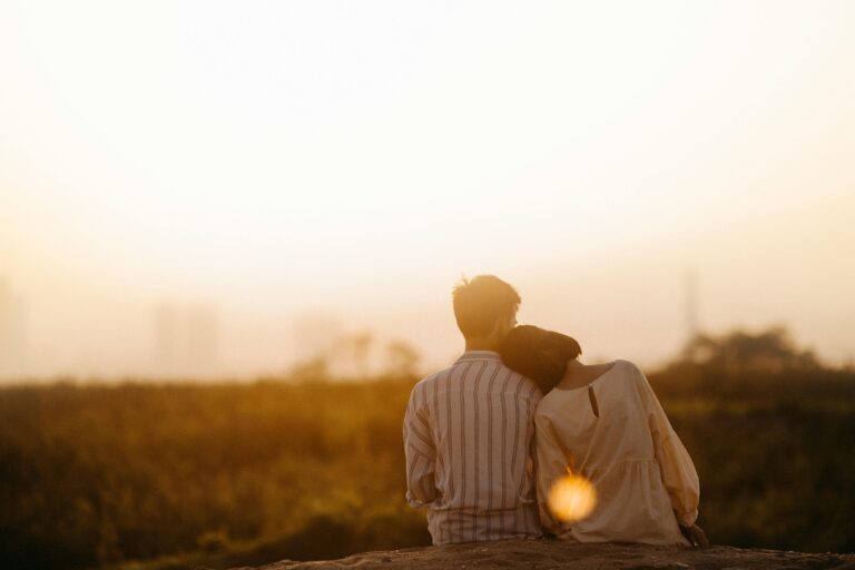 A couple sits closely together in a field at sunset, with the woman resting her head on the man's shoulder, creating a serene and intimate moment.