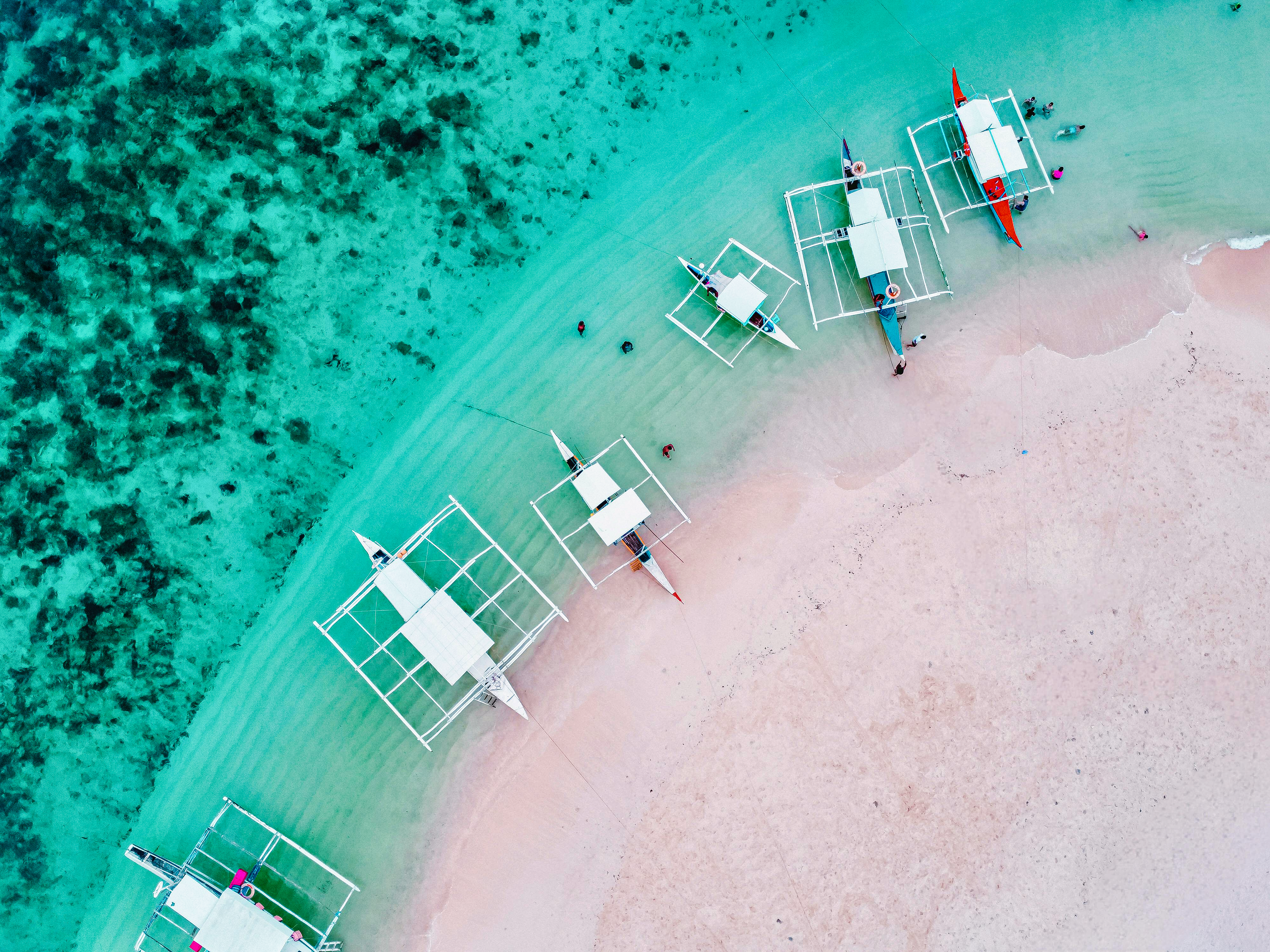 An aerial view of a beach with clear turquoise waters, showcasing traditional Filipino outrigger boats (bancas) lined up along the shoreline, with patches of coral visible beneath the surface and a few people enjoying the shallow water and sandy beach.