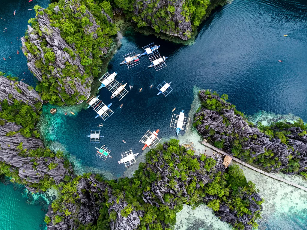 Aerial view of a tropical island in El Nido, Palawan with turquoise waters, limestone cliffs, white sand beach, palm trees, and traditional Filipino boats.