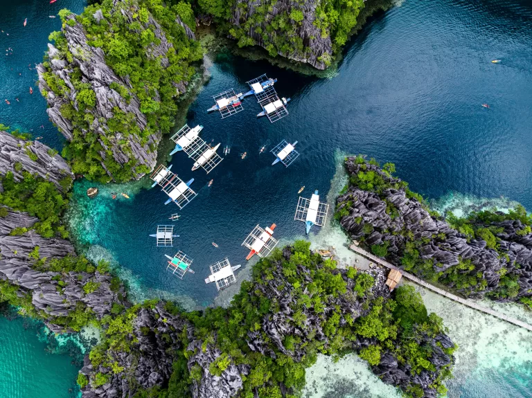 Aerial view of a tropical island in El Nido, Palawan with turquoise waters, limestone cliffs, white sand beach, palm trees, and traditional Filipino boats.