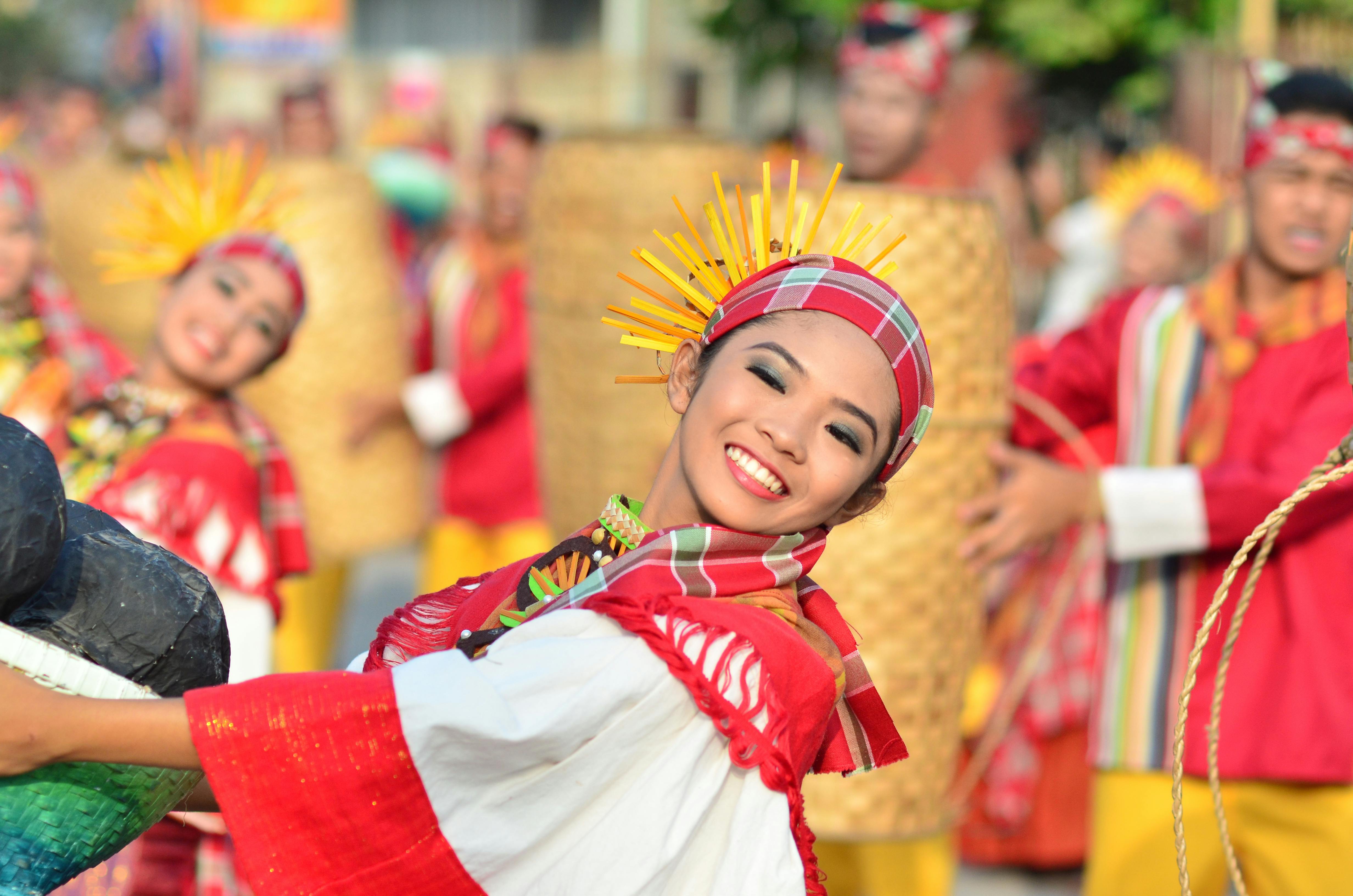 A woman dressed in traditional Filipino attire smiles brightly while dancing in a vibrant street parade, surrounded by other performers in colorful costumes.