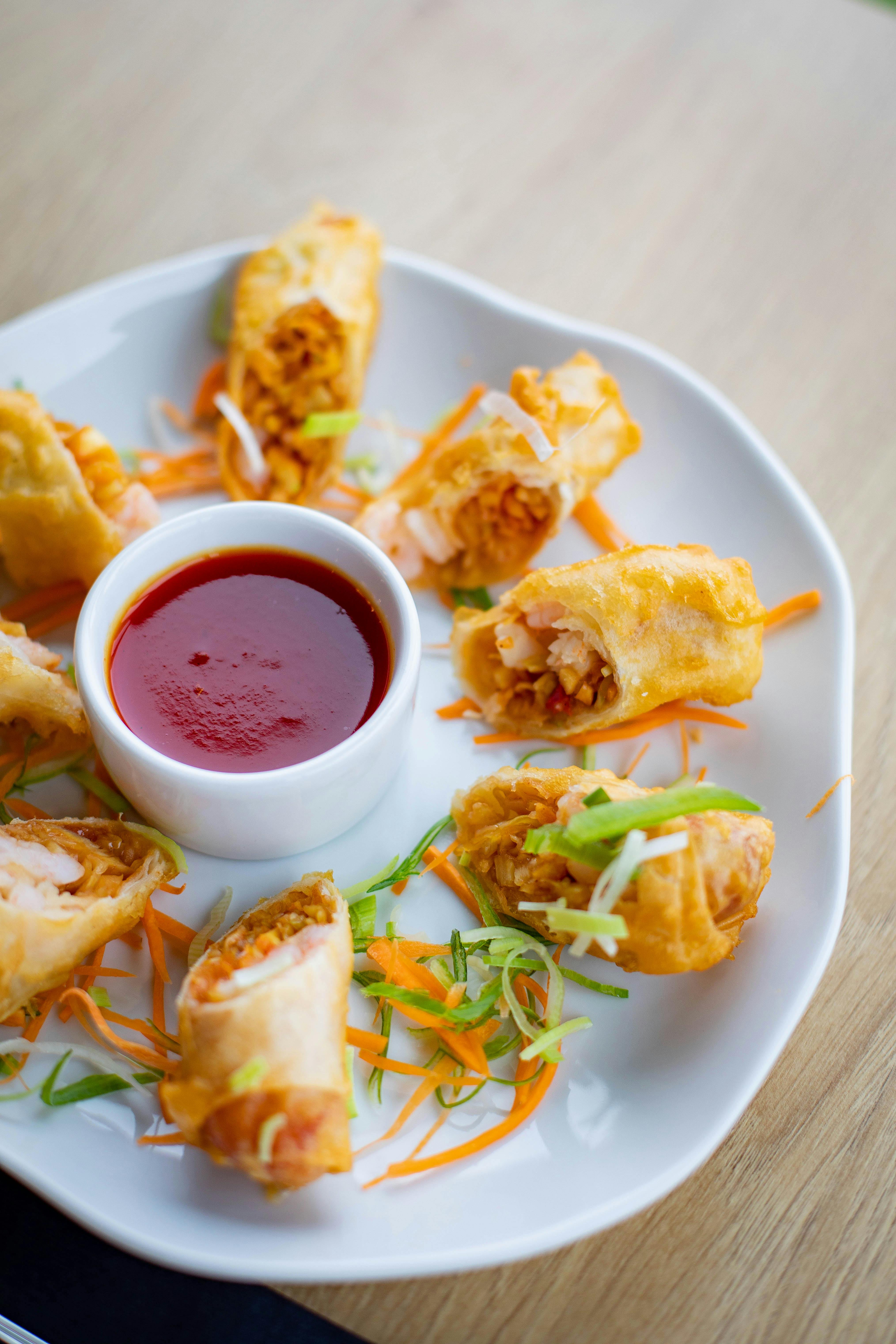 A plate of sliced Lumpiang Shanghai (Filipino spring rolls) arranged around a bowl of dipping sauce, garnished with shredded carrots and green onions.