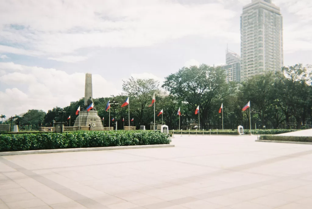 The Rizal Monument at Luneta Park in Manila, Philippines, surrounded by Philippine flags, trees, and a modern high-rise building in the background under a partly cloudy sky.