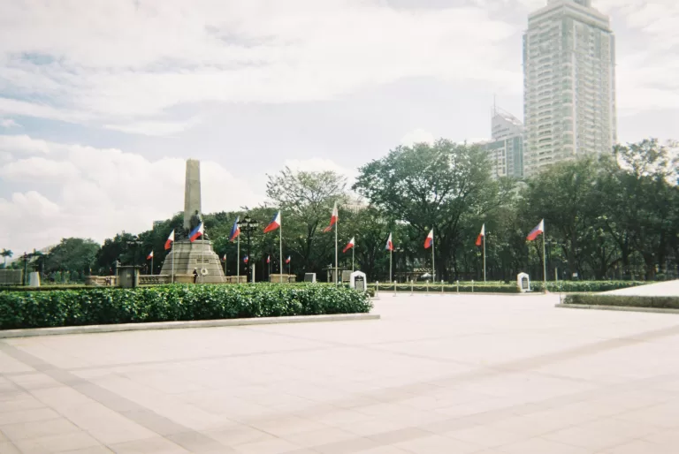 The Rizal Monument at Luneta Park in Manila, Philippines, surrounded by Philippine flags, trees, and a modern high-rise building in the background under a partly cloudy sky.