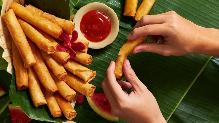 Hands holding a crispy Filipino lumpia spring roll above a banana leaf platter, surrounded by more golden fried rolls and a bowl of sweet chili dipping sauce.