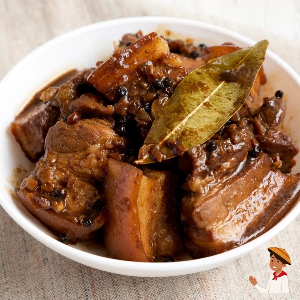 Filipino pork adobo in a white bowl with bay leaf and peppercorns.