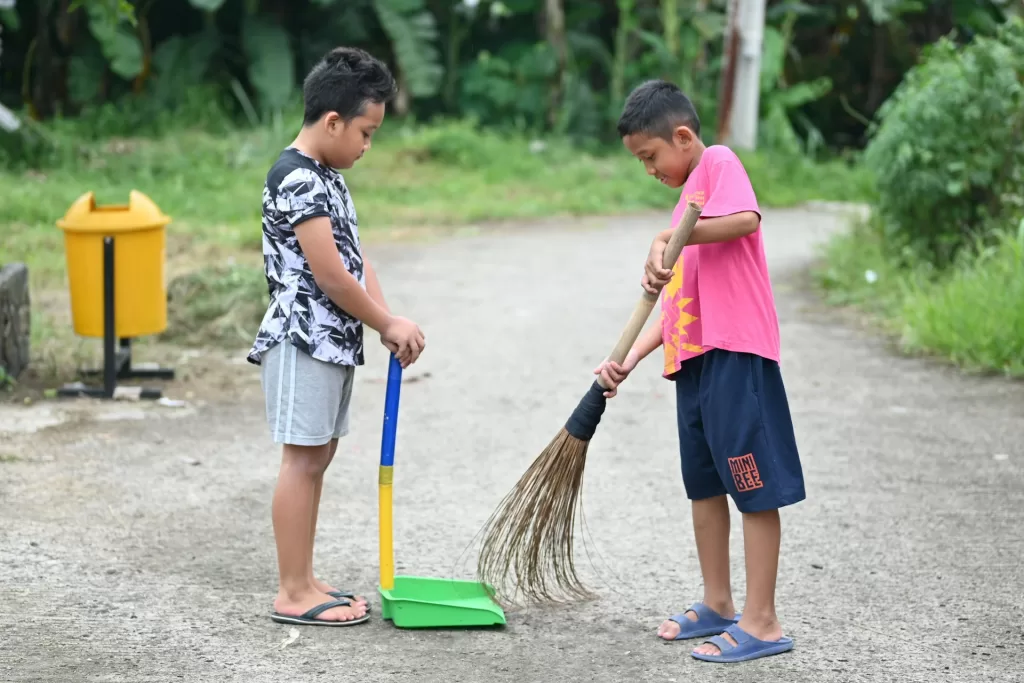 Two young Filipino boys sweeping a village street together with a walis tingting and dustpan, demonstrating the Filipino tradition of community cleanliness and shared responsibility or ‘bayanihan’ spirit.