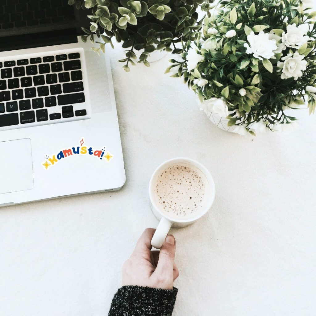 Laptop with colorful ‘Kamusta’ sticker beside coffee cup and plants